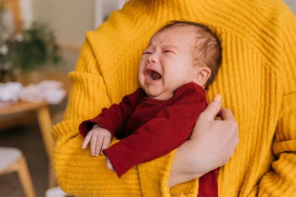 Tired mother holding a waking baby at night near crib in dim lighting